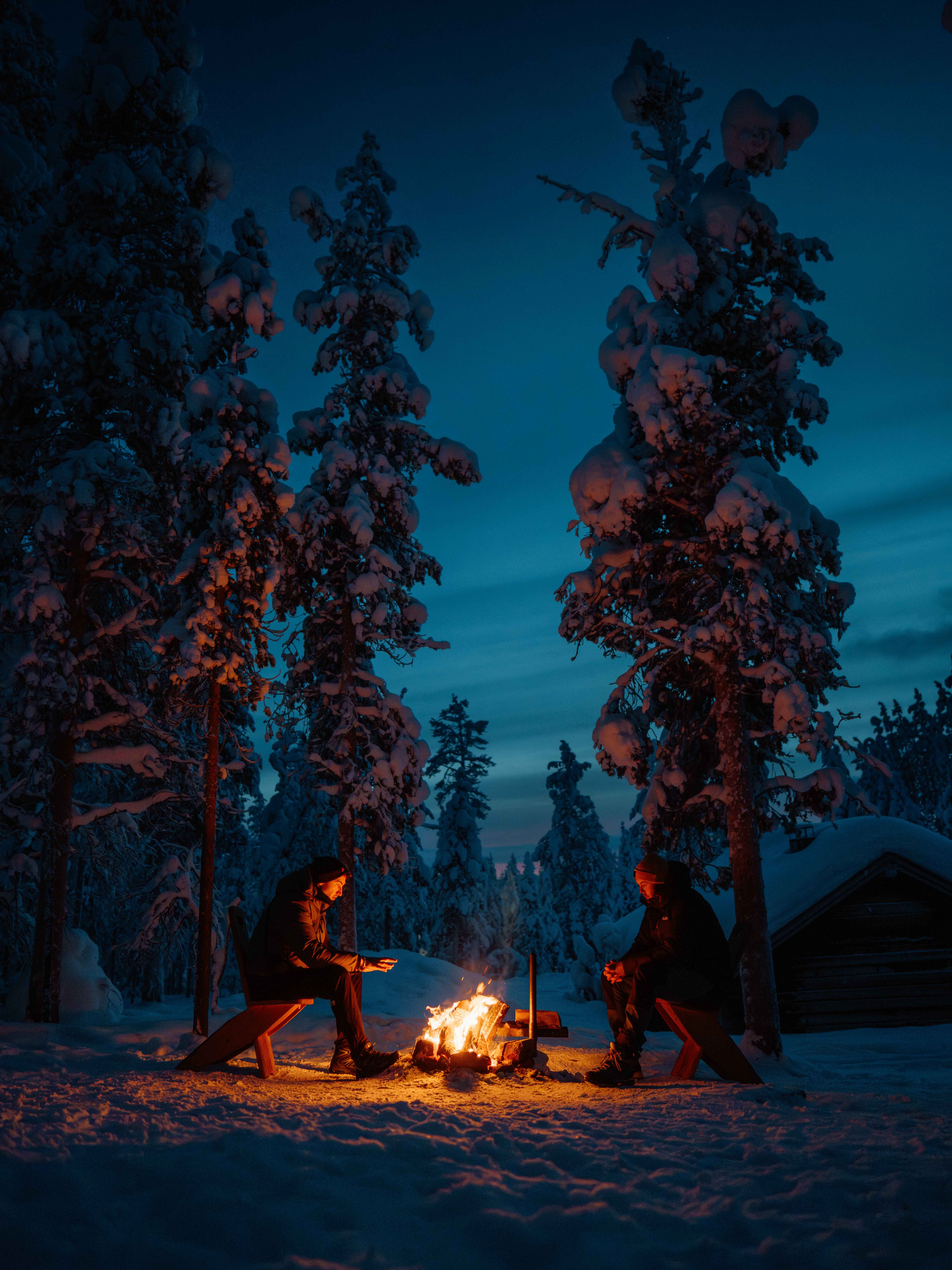 Two people sit around a campfire in a snowy forest at night